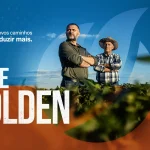 Banner publicitário horizontal com fundo dividido entre tons de azul e laranja, apresentando ao centro dois agricultores em pé em uma lavoura de soja sob céu limpo. Do lado esquerdo, destaca-se a frase "Existem novos caminhos para produzir mais." acima do texto em grandes letras brancas "VAI DE GOLDEN". No canto inferior direito, consta o logotipo da marca Golden Harvest com a assinatura "Uma marca Syngenta".